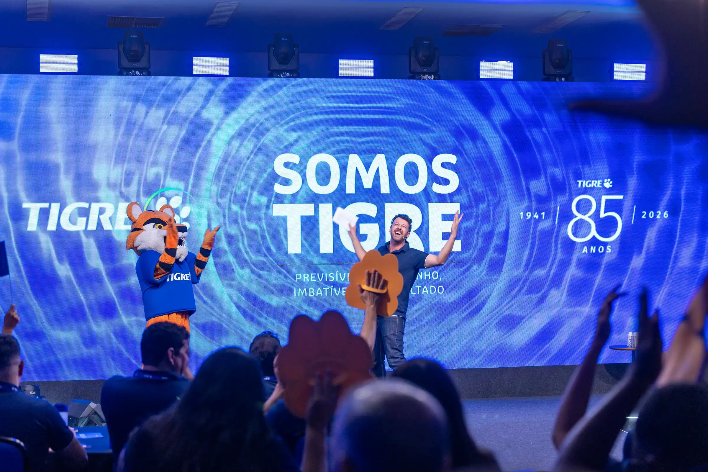 Tiago Lopes celebrando no palco com o público de braços erguidos em um evento corporativo