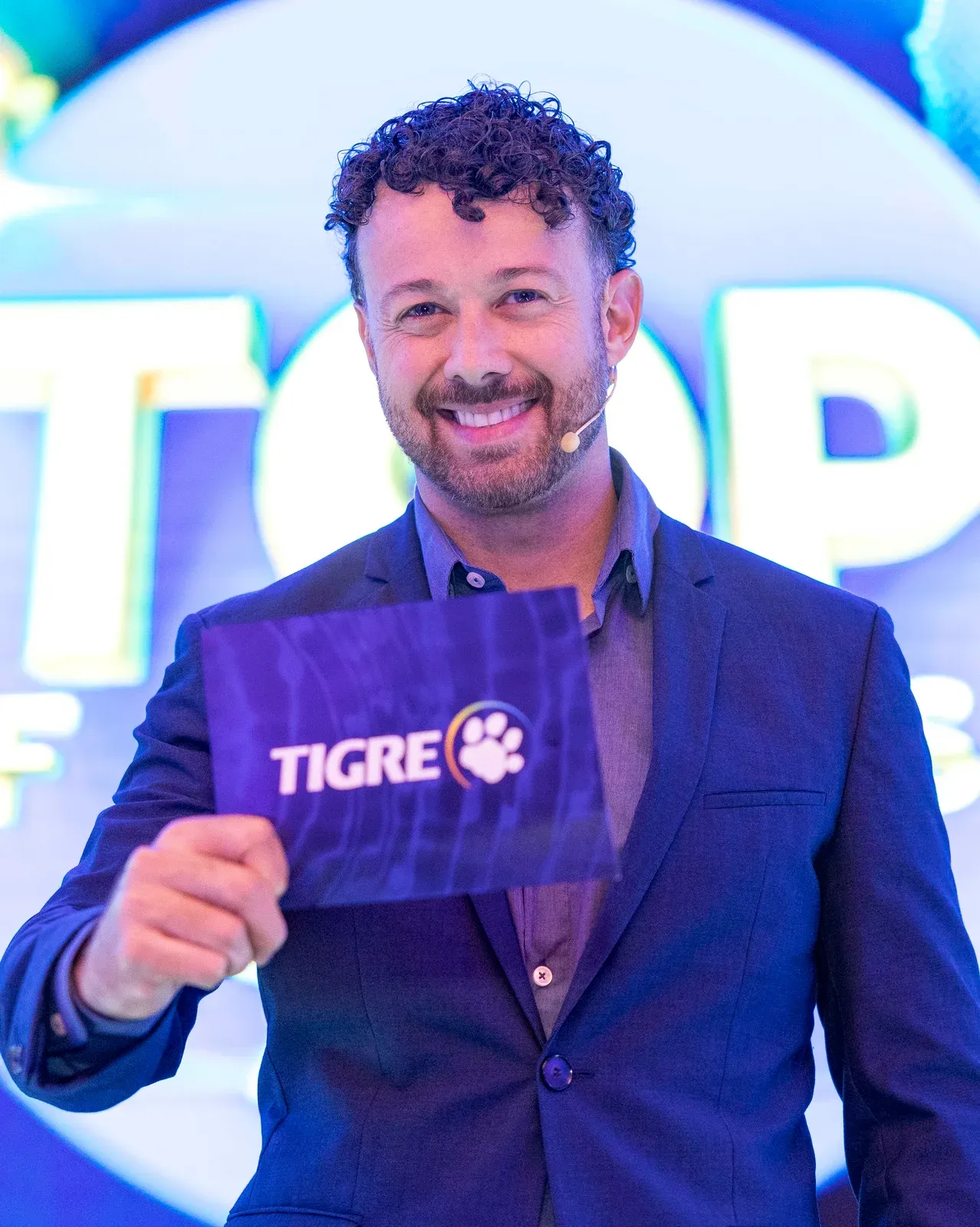 Tiago Lopes sorrindo no palco como mestre de cerimônias de uma premiação corporativa